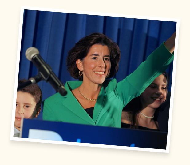 A photograph of Gina Raimondo standing a podium, smiling, and raising her arm.